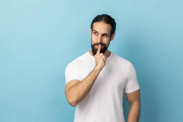 Shh, it\'s big secret. Portrait of man with beard wearing white T-shirt smiling, showing gesture secret sign with finger near his lips. Indoor studio shot isolated on blue background.