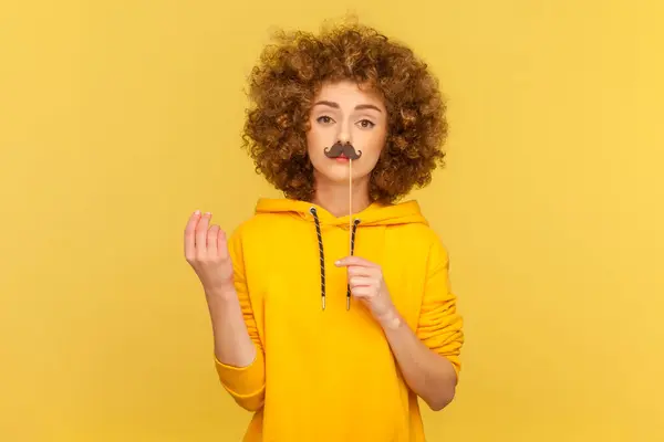 Portrait of funny playful woman with Afro hairstyle having fun on party with party props, holding paper mustache, wearing casual style hoodie. Indoor studio shot isolated on yellow background.