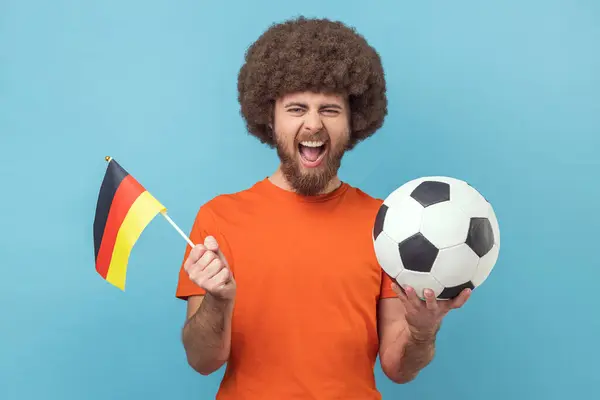 一个有着非洲式发型的疯狂男子的画像 他拿着德国国旗和足球的黑白经典球 观看着比赛 欢呼着 室内拍摄被蓝色背景隔离 — 图库照片