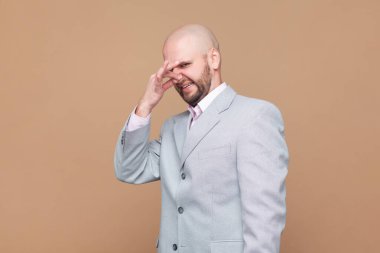 Tiksinerek yüzünü buruşturan kel sakallı adamın portresi, nefesini tutan, kötü kokmaması için parmaklarıyla burnunu çimdikleyen, berbat kokan, gri ceket giyen. Kahverengi arka planda kapalı stüdyo çekimleri.