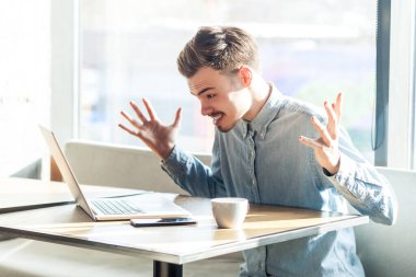 Sinirli, sakallı genç bir adamın portresi kafede oturmuş bilgisayarla tartışıyor, kot ceket giyiyor. Büyük pencereye karşı kapalı çekim.