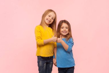 Gülümseyen iki küçük kızın baş parmaklarını gösterip, jest yaparak mutluluğu ifade etmesinin portresi. Pembe arka planda kapalı stüdyo çekimleri.