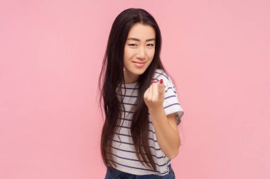 Portrait of playful woman with long brunette hair showing beckoning gesture saying come here, wearing striped T-shirt. Indoor studio shot isolated on pink background. clipart
