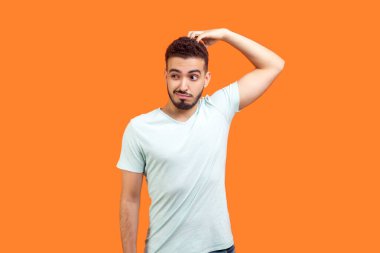 Düşünceli, sakallı, tişört giyen, başını ovuşturup başka tarafa bakmayı düşünen genç bir adamın portresi. Turuncu arka planda kapalı stüdyo çekimleri.