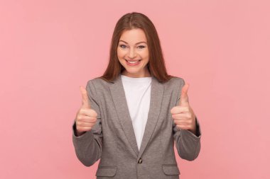Kahverengi saçlı, gülümseyen bir kadının portresi başparmağını kaldırıyor, servisi seviyor, mutluluğu ifade ediyor, takım elbise giyiyor. Pembe arka planda kapalı stüdyo çekimleri.