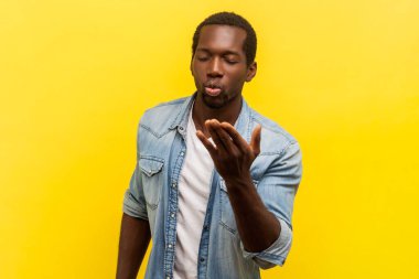 Romantik flörtçü adamın portresi. Kapalı gözlerle dikilmiş, havada öpüşen, aşkını ifade eden, kot gömleği giyen. Sarı arka planda kapalı stüdyo çekimleri.