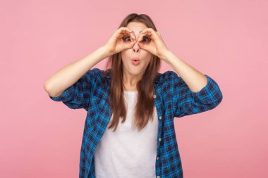 Şaşırmış kahverengi saçlı kadının portresi. Kameraya bakıyor. Dürbünlü bir yüz ve kareli gömlek giyiyor. Pembe arka planda kapalı stüdyo çekimleri.