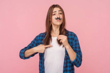 Kahverengi saçlı komik bir kadının portresi. Elinde kağıt bıyıkla dikilip kameraya bakıyor, kareli gömlek giyiyor. Pembe arka planda kapalı stüdyo çekimleri.