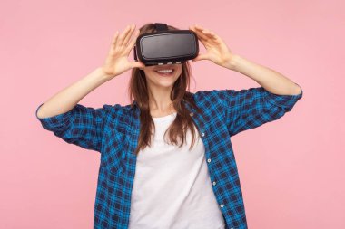VR kulaklıklı kahverengi saçlı kadın portresi, yüzünde gülümsemeyle sanal gerçeklik oyunu oynuyor, yenilikçi teknoloji, kareli gömlek giyiyor. Pembe arka planda kapalı stüdyo çekimleri.