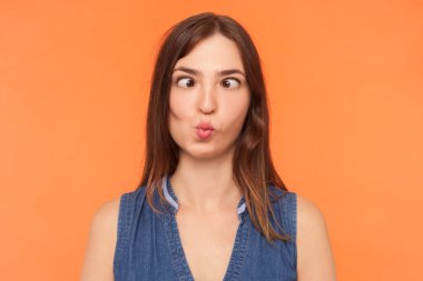 Kot elbise giyen, gözlerini çaprazlayan, balık dudaklı, suratını buruşturarak eğlenen çocuksu, komik esmer kadının portresi. Turuncu arka planda kapalı stüdyo çekimleri.