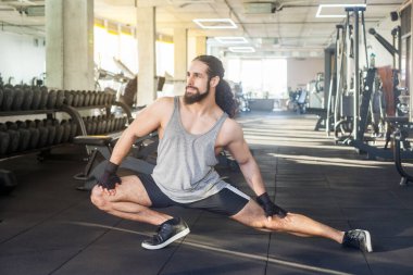 Esnek, kıvırcık saçlı spor giysili spor kıyafetleri giyen spor salonunda egzersiz yapmadan önce bacaklarını ısıtan spor salonuna gidip yan bakan kaslı bir adamın portresi. Kapalı çekim.