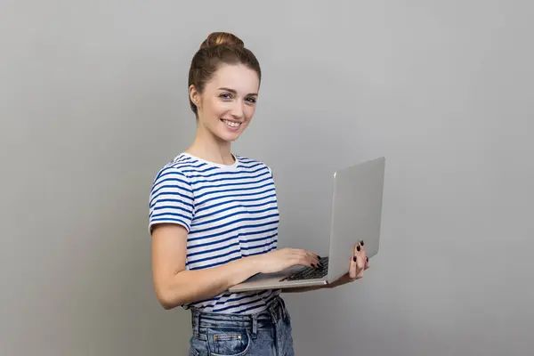 穿着条纹T恤的快乐女性在笔记本电脑上愉快地工作的侧面视图 喜欢自由职业 看着相机 在灰色背景下被隔离的室内拍摄 免版税图库图片