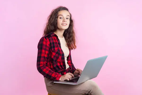 信頼できる十代の少女の肖像画 赤いチェッカーのシャツの重い髪 ラップトップのリモート作業距離の仕事に座っています ピンクの背景に隔離された屋内スタジオショット ロイヤリティフリーのストック写真