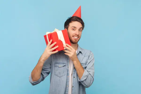 パーティーコーンのポジティブなハンサムな男の笑顔の肖像画は プレゼントボックスを包んで デニムシャツを着用して誕生日を祝う ブルーの背景に隔離された屋内ショット ロイヤリティフリーのストック写真