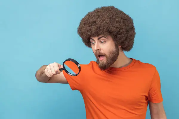 穿着橙色T恤的黑发男子的画像 透过放大镜 探索犯罪现场 室内拍摄被蓝色背景隔离 免版税图库照片