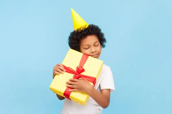 可爱的小男孩的画像 卷曲的头发 戴着黄色的派对锥 站在那里抱着生日礼物 闭着眼睛享受着 室内拍摄被蓝色背景隔离 图库图片