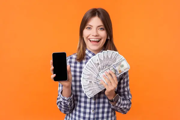 一个面带微笑 积极向上 一头棕色头发的女人的画像 她穿着格子衬衫 带着空的显示屏和美元钞票 展示着智能手机 在橙色背景下被隔离的室内拍摄 图库图片