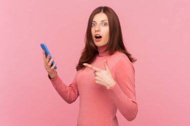 Kahverengi saçlı, cep telefonu kullanan, ekranda büyük gözlerle kameraya bakan, şok içerikli, balıkçı yaka giyen şaşkın bir kadın. Pembe arka planda kapalı stüdyo çekimleri