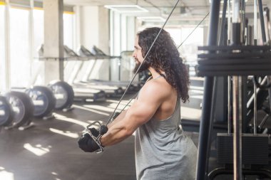 Sağlıklı bir yaşam tarzı. Spor salonunda uzun kıvırcık saçlı sporcu portresi ayakta duran ve ağırlık kaldıran, bisepsler için egzersiz yapan, antrenman için simülatör kullanan. Kapalı çekim.