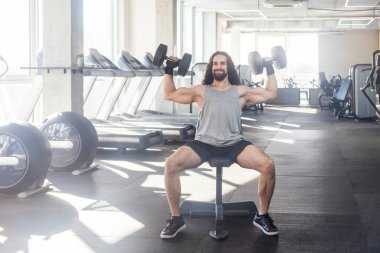 Uzun kıvırcık saçlı sporcu özgüvenli bir adamın portresi. Spor salonunda çalışıyor, ağırlık kaldırıyor ve kollarını kaldırıyor. Pazılar ve trisepsler için egzersiz yapıyor. Kapalı çekim.