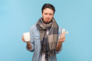 Sıcak atkıya sarılmış, boğazı ağrıyan, elinde çay ve elinde haplarla kot gömlek giyen sağlıksız, hasta, hasta bir adamın portresi. Mavi arkaplanda kapalı çekim.