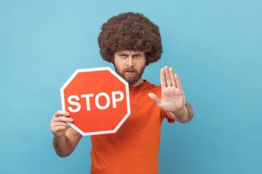 Turuncu tişörtlü Afro saçlı bir adamın portresi. Durma hareketi gösteriyor. Trafik levhası tutuyor. Mavi arka planda kapalı stüdyo çekimi.