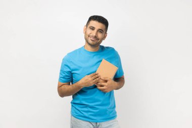 Mavi tişört giyen, elinde kitapla kameraya bakıp gülümseyen neşeli ve pozitif gülümseyen bir adamın portresi. Gri arka planda izole edilmiş kapalı stüdyo görüntüleri.