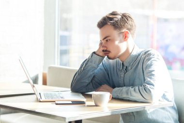 Kafede oturup dizüstü bilgisayarda çalışan, Webinar seyreden, kot ceket giyen ciddi konsantre sakallı genç bir adamın portresi. Büyük pencereye karşı kapalı çekim.
