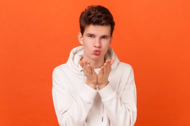 Beyaz kapüşonlu genç adam portresi hava öpücüğü veriyor, avuç içini ağzına doğru katlıyor, kameraya bakarak mwah coquettish gönderiyor. Turuncu arka planda kapalı stüdyo çekimleri.