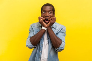 Şok olmuş, tırnaklarını yiyen kameraya bakan gergin bir adamın portresi korkunç bir şey görüyor, sıradan bir kot gömlek giyiyor. Sarı arka planda kapalı stüdyo çekimleri.