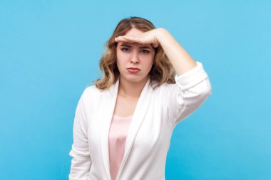 Dalgalı saçlı, ciddi, meraklı sarışın kadın portresi, elini alnının yakınına uzatıyor. Görmeye çalışıyor, beyaz gömlek giyiyor. Mavi arka planda kapalı stüdyo çekimi.