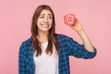 Kahverengi saçlı komik bir kadının portresi, elinde donut tutarak bakışlarını kaçırıyor, yiyor ya da yemiyor, dişlerini sıkmaya devam ediyor, kareli gömlek giyiyor. Pembe arka planda kapalı stüdyo çekimleri.