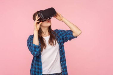 VR kulaklıklı konsantre kahverengi saçlı kadın portresi, sanal gerçeklik oyunu oynuyor, yenilikçi teknoloji, kareli gömlek giyiyor. Pembe arka planda kapalı stüdyo çekimleri.