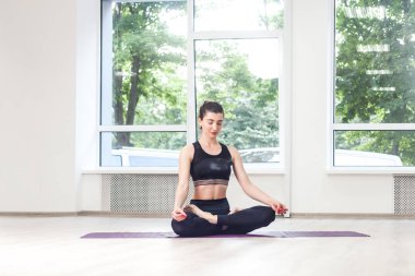 Yoga yapan, Lotus pozu veren, siyah pantolon ve üst giyen, meditasyon yapan, sakin, gerçekçi genç bir kadının portresi. Spor salonunda, büyük pencerelerin yanında..