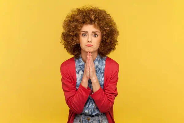 stock image Portrait of pleading woman with Afro hairstyle holding hands folded in praying gesture, asking for help, pressing palms together. Indoor studio shot isolated on yellow background.