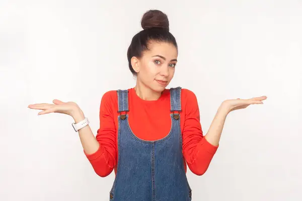 stock image Portrait of uncertain puzzled woman with hair bun shrugging shoulders, dosen't know answer the question, wearing denim overalls. Indoor studio shot isolated on white background
