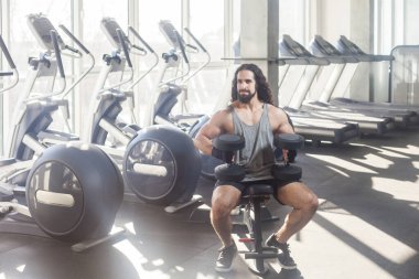 Uzun kıvırcık saçlı, spor salonunda çalışan, bankta oturan, dambılları tutan, pazılar ve pazılar için kolunu kaldıran ve başka yöne bakan yakışıklı, güçlü bir adamın portresi. Kapalı çekim.