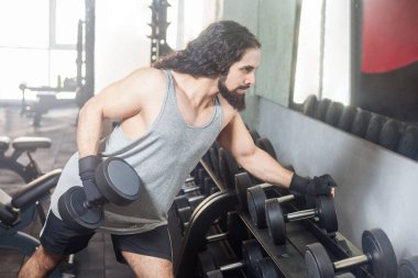 Uzun kıvırcık saçlı spor kıyafetleri giyen spor spor spor salonunda spor yapan kaslı genç bir adamın portresi. Kol ve kol kası egzersizleri için halterler alıyor..