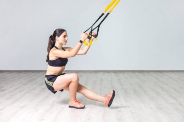 Spor kıyafetleri giyen, spor giyerek spor salonundaki spor kemerleriyle çömelmiş vücut egzersizi için trx kullanan güzel bir kadının yan görüntüsü. Beyaz duvar arkaplanına karşı kapalı çekim