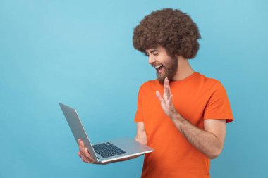 Afro saçlı, turuncu tişörtlü, dizüstü bilgisayarlı, video görüşmeli, el sallayan, selamlayan dost canlısı bir adamın portresi. Mavi arka planda kapalı stüdyo çekimi.