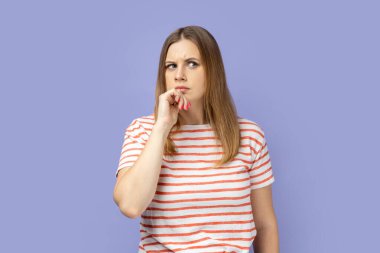 Kafası karışık sarışın kadın çizgili tişört giyiyor. Düşüncelere dalmış, şaşkın, ciddi şüpheli yüz ifadesiyle zor kararlar veriyor. Mor arka planda izole edilmiş kapalı stüdyo çekimi.