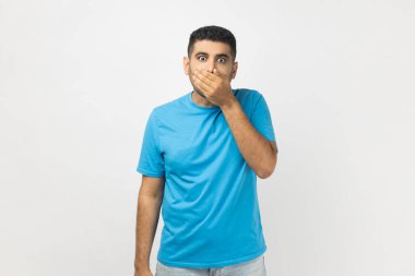 Mavi tişört giyen, ağzı avuç içiyle kaplanmış ve kimseye söylemeyeceğimi söyleyen şaşkın ve tıraşsız bir adamın portresi. Gri arka planda izole edilmiş kapalı stüdyo görüntüleri.