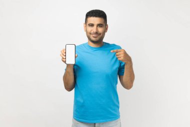 Mavi tişört giyen, mutlu, sevimli, tıraşsız bir adamın portresi boş akıllı telefon ekranını göstererek kameraya bakıyor. Gri arka planda izole edilmiş kapalı stüdyo görüntüleri.