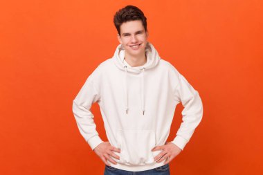 Beyaz kapüşonlu giyen, elleri kalçasında duran ve dişlek bir gülümsemeyle kameraya bakan neşeli, kendine güvenen genç bir adamın portresi. Turuncu arka planda kapalı stüdyo çekimleri.