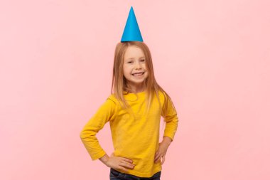 Mavi parti konisindeki sevimli sarışın kız portresi ellerini kalçasında tutuyor, doğum günü partisinde eğleniyor, sarı kazak giyiyor. Pembe arka planda kapalı stüdyo çekimleri.