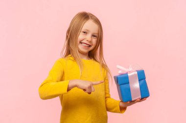 Gülümseyen iyimser küçük kızın portresi mavi hediye kutusunu tutan, dişlek tebessümle kameraya bakan, sarı kazak giyen. Pembe arka planda kapalı stüdyo çekimleri.