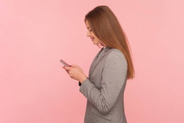 Kahverengi saçlı, gülümseyen bir kadının portresi. Akıllı telefon kullanıyor, arkadaşlarıyla sohbet ediyor, internette iletişim kuruyor, takım elbise giyiyor. Pembe arka planda kapalı stüdyo çekimleri.