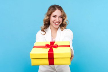 Gülümseyen, dalgalı saçlı, sarı ambalajlı hediye kutusu taşıyan, beyaz gömlek giyen neşeli sarışın kadının portresi. Mavi arka planda kapalı stüdyo çekimi.