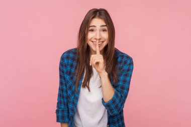 Gülümseyen, kahverengi saçlı, ağzı kapalı bir kadın kameraya olumlu bir ifadeyle bakıyor, kareli gömlek giyiyor. Pembe arka planda kapalı stüdyo çekimleri.