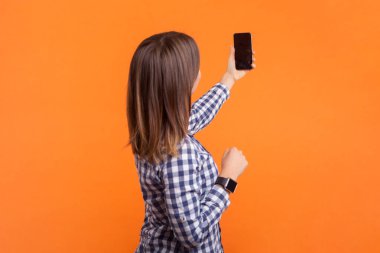 Kahverengi saçlı, akıllı telefonlu, selfie yapan, canlı yayın yapan, kareli gömlek giyen bir kadın. Turuncu arka planda kapalı stüdyo çekimleri